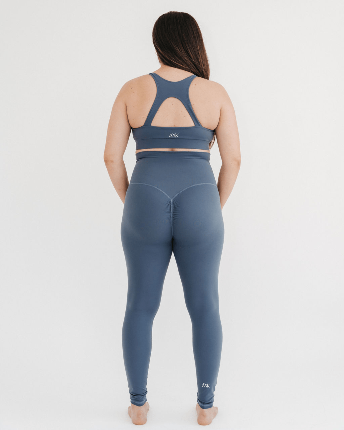 Pregnant woman wearing premium blue maternity leggings with a flattering scrunch butt and matching high-impact, racerback nursing sports bra, standing against a white background. Premium maternity athletic and activewear by AXK.