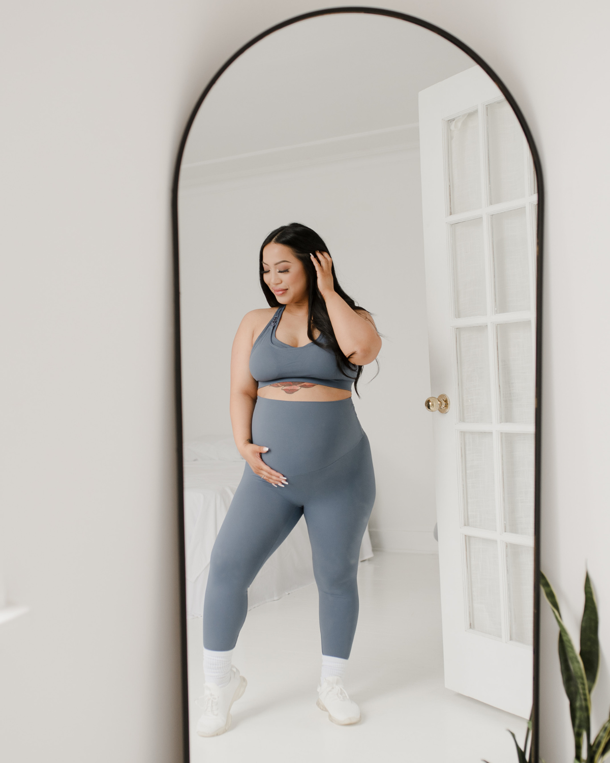 Pregnant woman wearing premium blue maternity leggings with a flattering scrunch butt and matching high-impact, racerback nursing sports bra, standing in front of a full-length mirror in a bright white room. Premium maternity athletic and activewear by AXK.