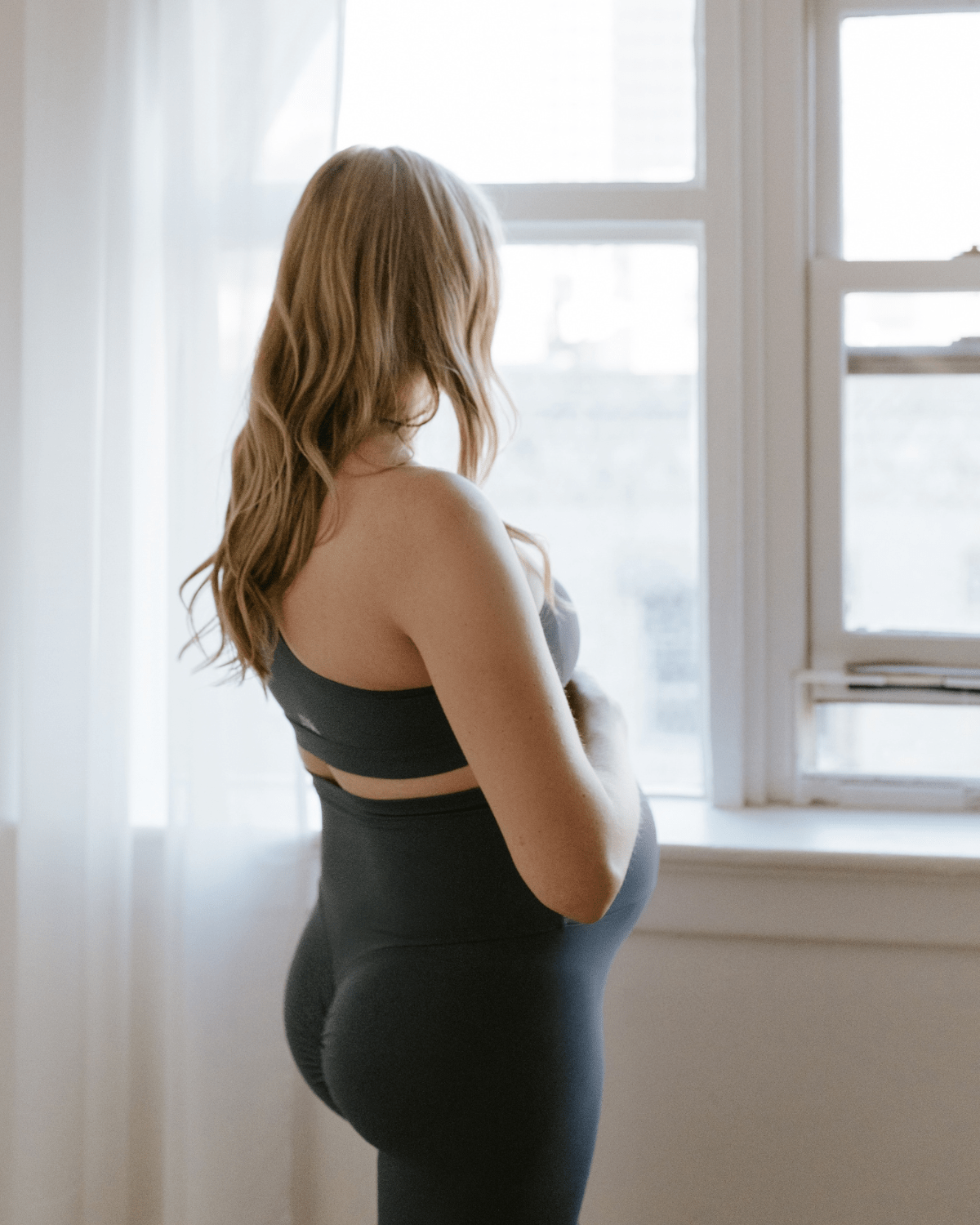 Pregnant woman wearing premium blue maternity leggings with a flattering scrunch butt and matching one-shoulder, nursing sports bra, standing in profile near a bright window. Premium maternity athletic and activewear by AXK.