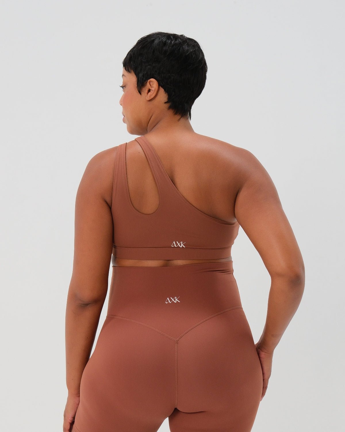 Pregnant woman wearing a brown one-shoulder maternity and nursing sports bra with matching high-waist leggings, standing against a white background. Premium maternity athletic and activewear by AXK - high quality petite, tall, and plus size maternity.