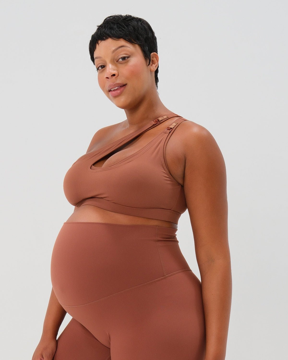 Pregnant woman wearing a brown one-shoulder maternity and nursing sports bra with matching high-waist leggings, standing against a white background. Premium maternity athletic and activewear by AXK - high quality petite, tall, and plus size maternity.