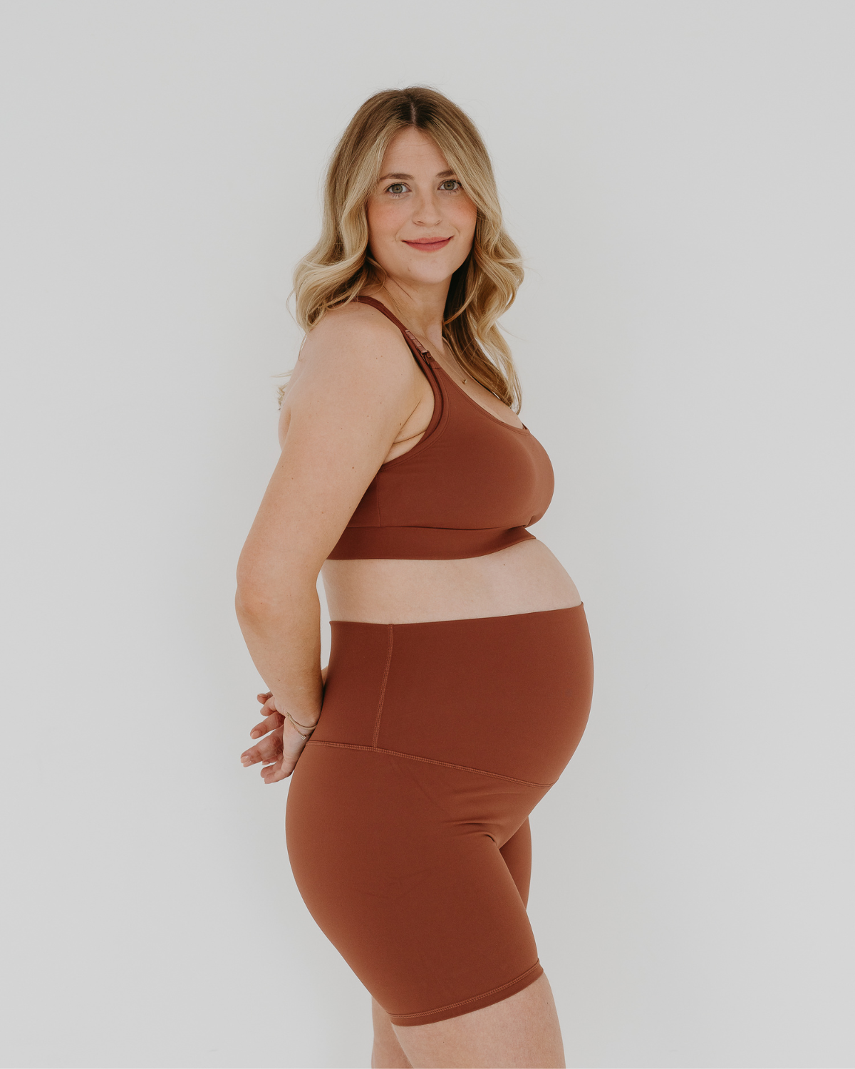 Pregnant woman wearing premium brown AXK maternity bike shorts with a supportive high waist and matching high-impact, racerback maternity and nursing sports bra, standing against a white background. Premium maternity athletic and activewear by AXK - high quality petite, tall, and plus size maternity.