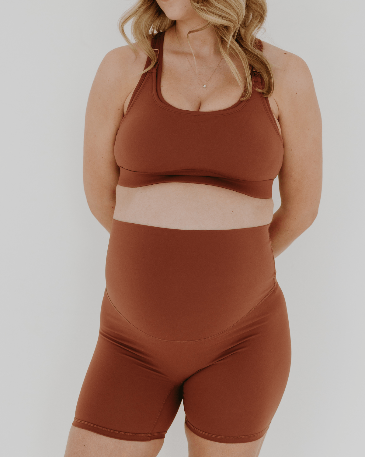 Pregnant woman wearing premium brown AXK maternity bike shorts with a supportive high waist and matching high-impact, racerback maternity and nursing sports bra, standing against a white background. Premium maternity athletic and activewear by AXK - high quality petite, tall, and plus size maternity.