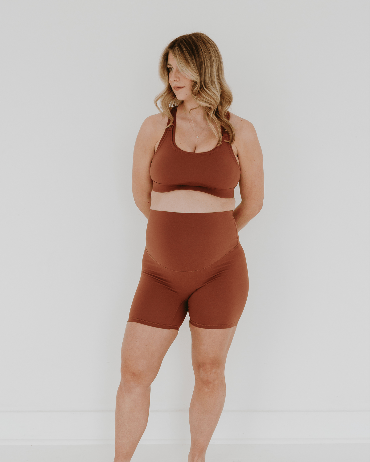 Pregnant woman wearing premium brown maternity bike shorts with a supportive high waist and matching high-impact, racerback maternity and nursing sports bra, standing against a white background. Premium maternity athletic and activewear by AXK - high quality petite, tall, and plus size maternity.