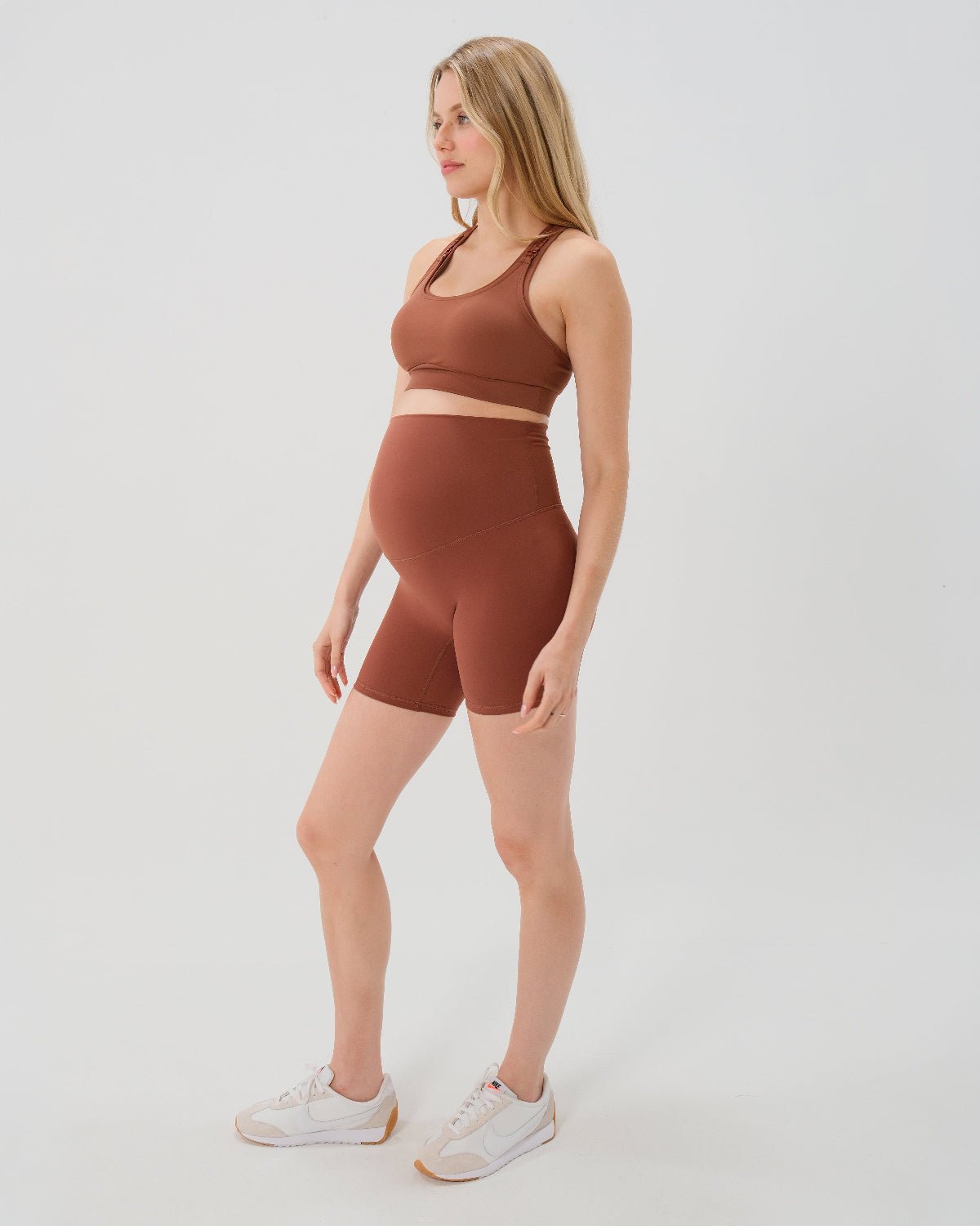 Pregnant woman wearing premium brown maternity bike shorts with a supportive high waist and matching high-impact, racerback maternity and nursing sports bra, standing against a white background. Premium maternity athletic and activewear by AXK - high quality petite, tall, and plus size maternity.