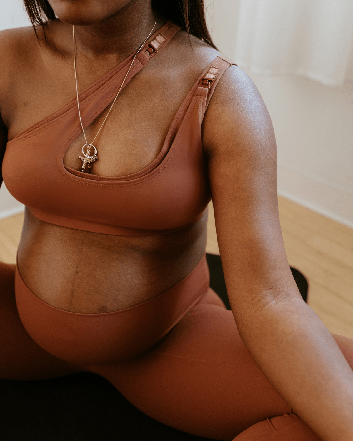 Close-up of a pregnant woman wearing a brown one-shoulder maternity and nursing sports bra with matching high-waist leggings, sitting cross-legged on a yoga mat indoors. Premium maternity activewear by AXK.