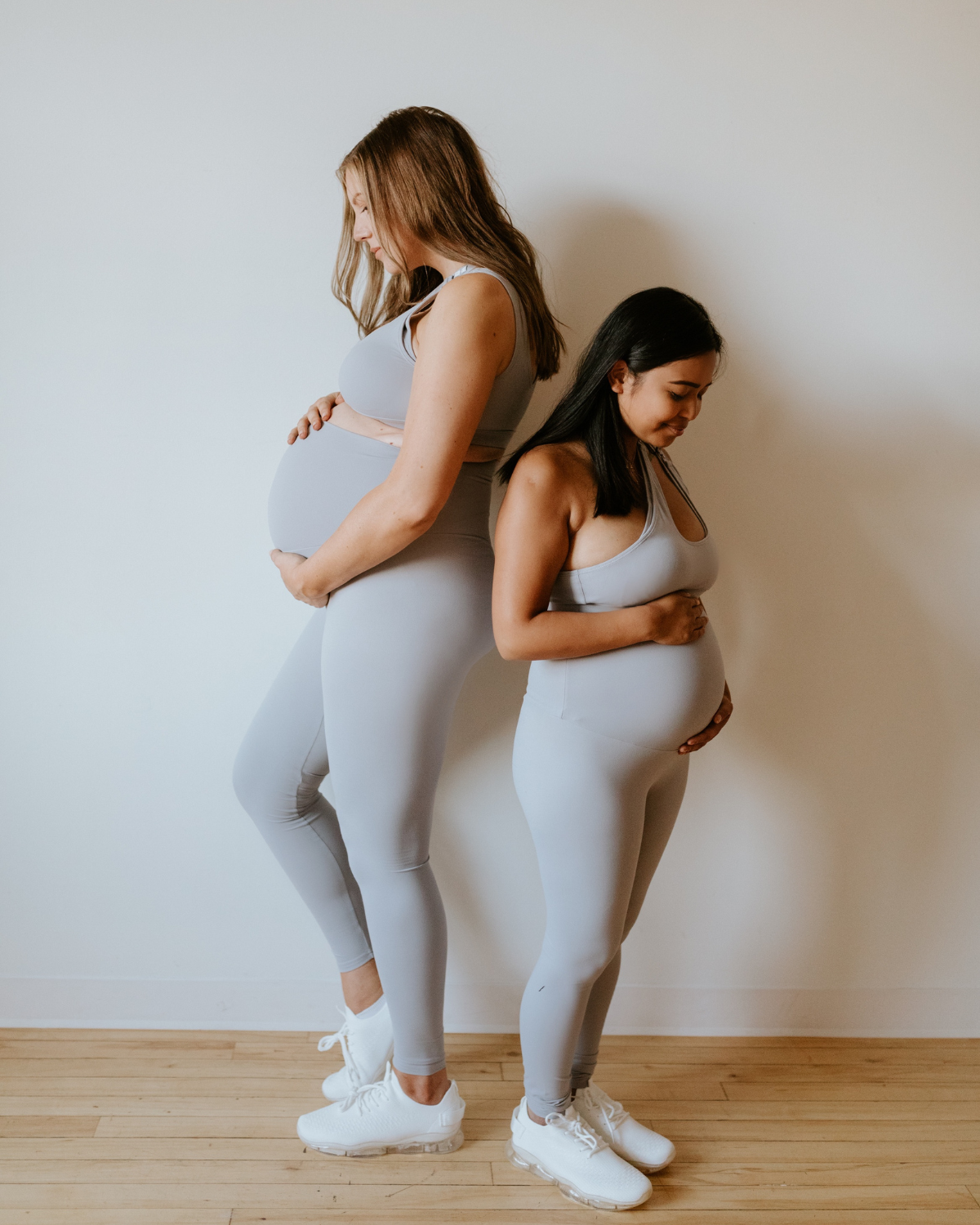 Two pregnant women wearing premium light grey maternity leggings with a flattering scrunch butt and matching one-shoulder, low-impact, nursing sports bras, standing side by side on a wood floor against a white wall. Premium maternity athletic and activewear by AXK.