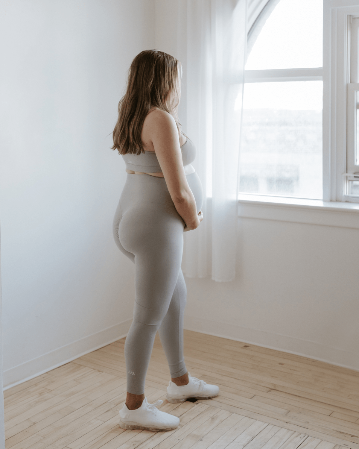 Pregnant woman wearing premium light grey maternity leggings with a flattering scrunch butt and matching one-shoulder, low-impact, nursing sports bra, standing in profile near a bright window. Premium maternity athletic and activewear by AXK.