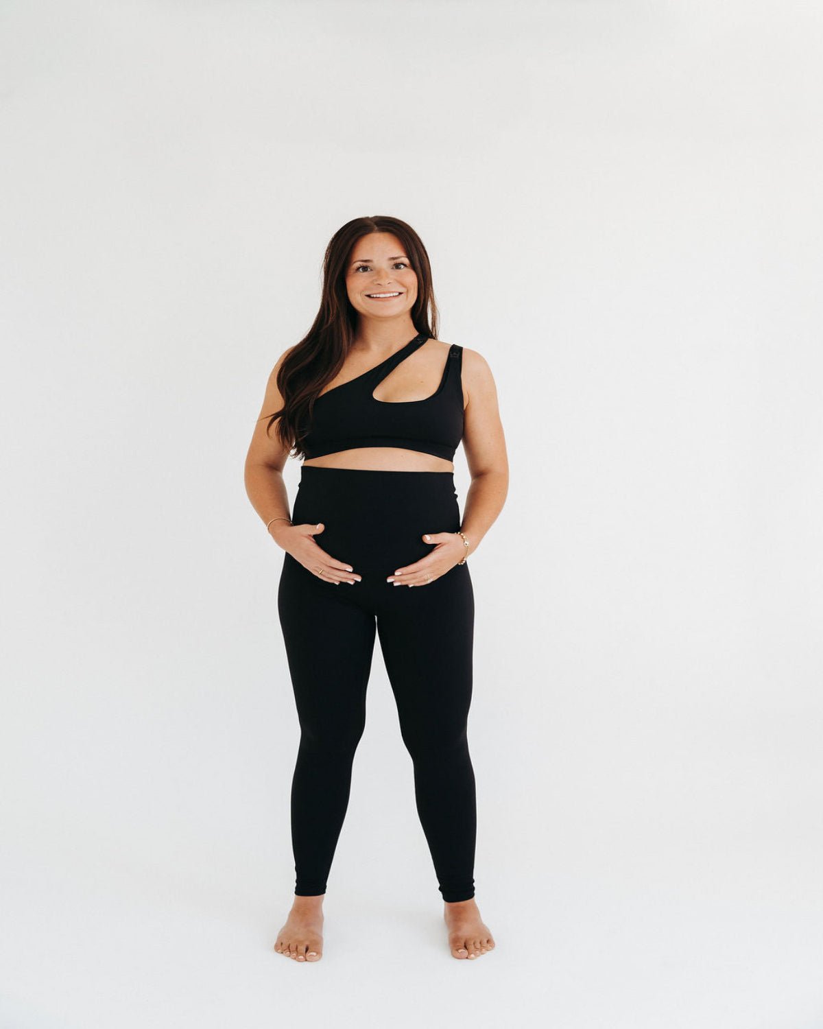 Pregnant woman wearing premium black maternity leggings with a flattering scrunch butt and matching high-impact, racerback nursing sports bra, standing against a white background. Premium maternity athletic and activewear by AXK.