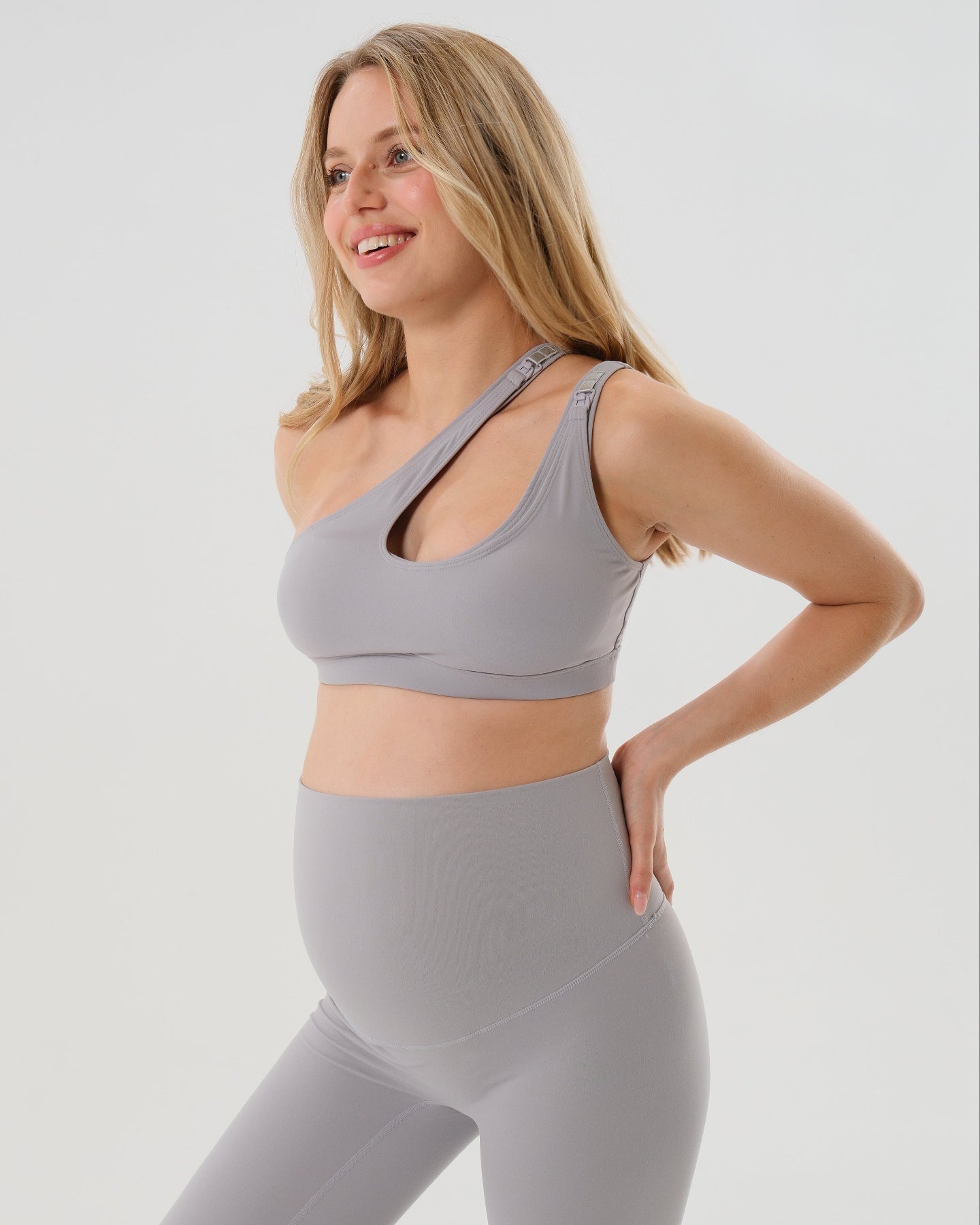 Pregnant woman in a one-shoulder light gray maternity and nursing sports bra, wearing matching light gray maternity leggings. Best selling 2026 premium maternity athletic and activewear by AXK, offered in regular and plus sizes, with glowing 5 star reviews. 