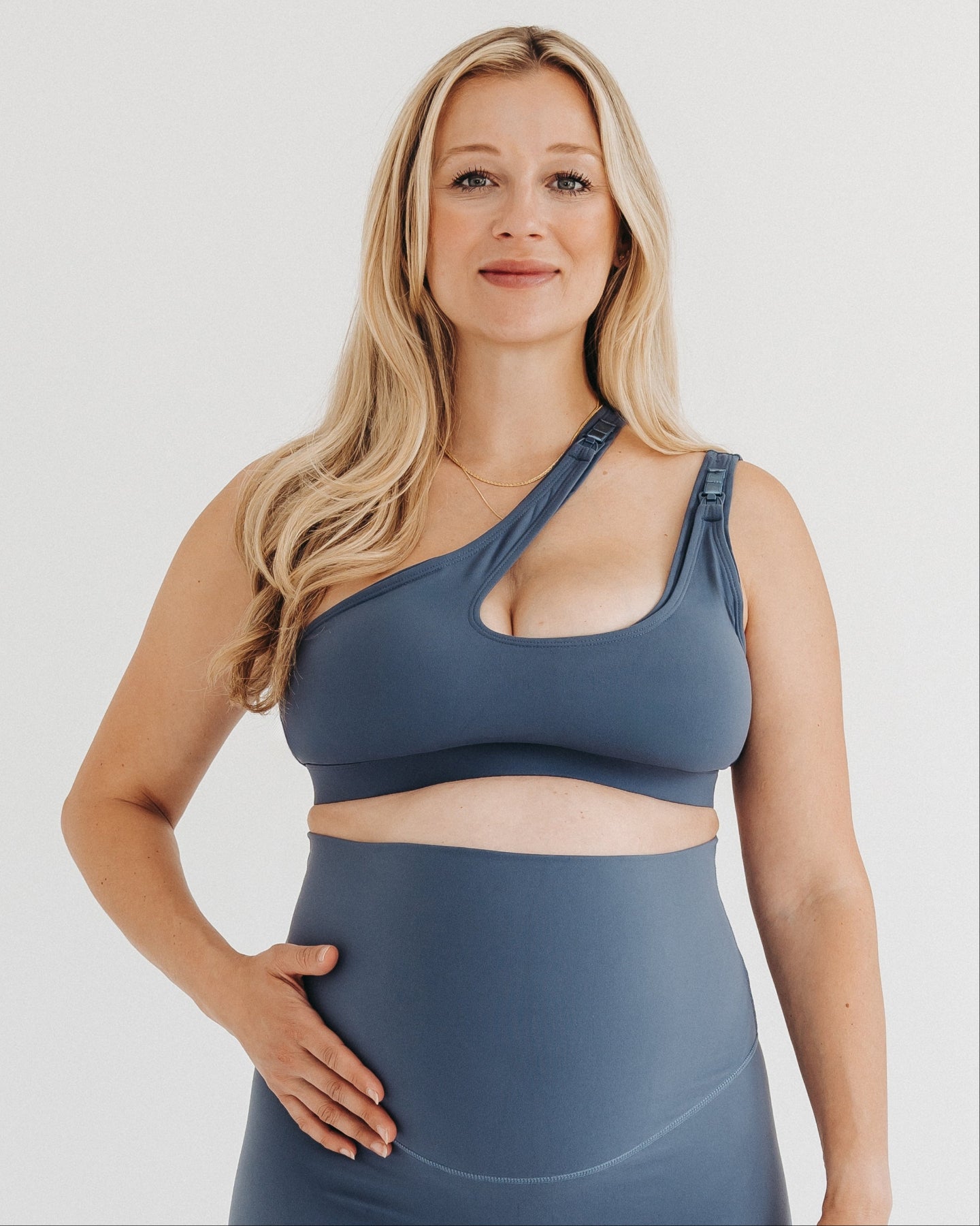 Pregnant woman in a one-shoulder blue maternity and nursing sports bra, wearing matching blue maternity leggings. Best selling 2026 premium maternity athletic and activewear by AXK, offered in regular and plus sizes, with glowing 5 star reviews. 