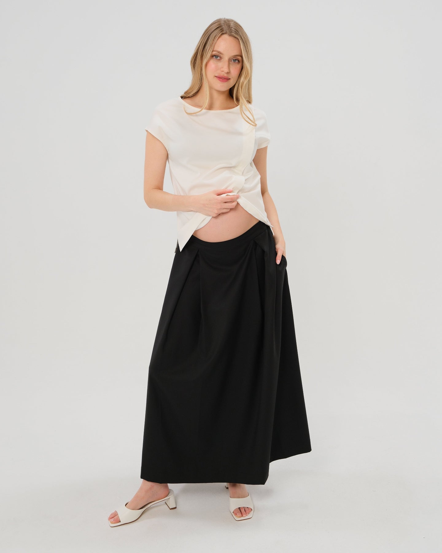 Pregnant woman wearing a black pleated designer maternity maxi maternity skirt in lightweight tropical wool by MARION. This luxury skirt can be worn over or under your belly bump. It fastens with a zip, and features deep functional pockets and a flexible elastic waistband for comfort. This refined, timeless style epitomizes quiet luxury. Perfect for maternity work wear, wedding guest dress, occasion wear, pregnancy street style, and baby shower outfits. Petite and tall friendly.