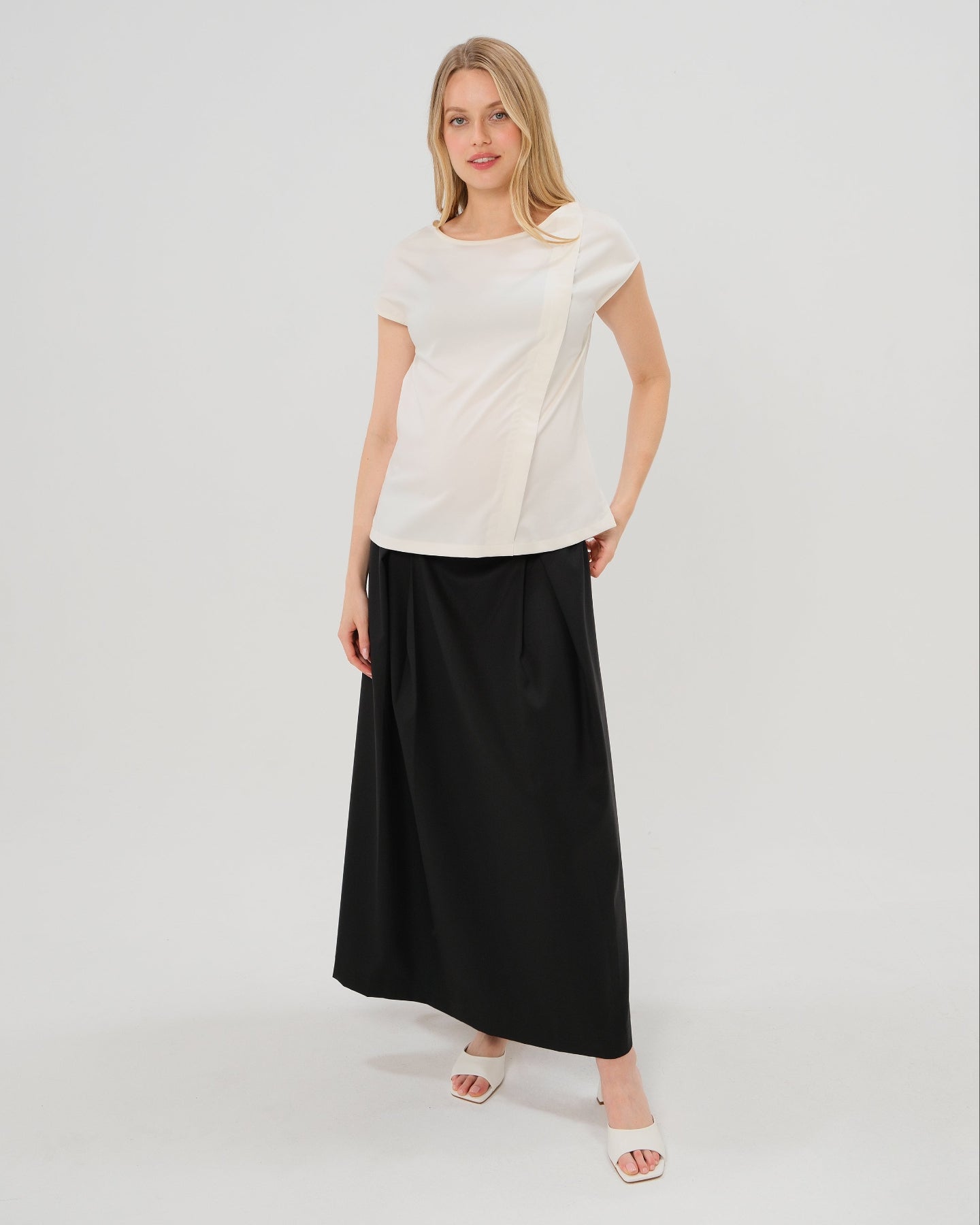 Pregnant woman wearing a black pleated designer maternity maxi maternity skirt in lightweight tropical wool by MARION. This luxury skirt can be worn over or under your belly bump. It fastens with a zip, and features deep functional pockets and a flexible elastic waistband for comfort. This refined, timeless style epitomizes quiet luxury. Perfect for maternity work wear, wedding guest dress, occasion wear, pregnancy street style, and baby shower outfits. Petite and tall friendly.