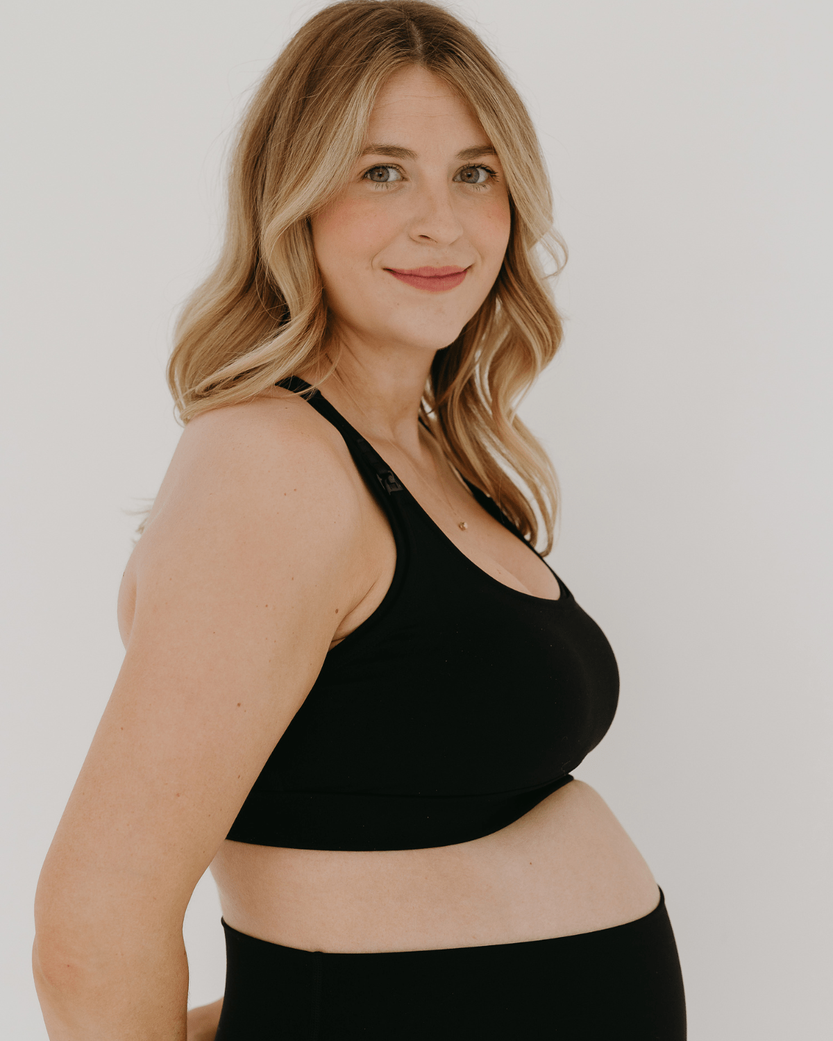 Side view of a pregnant woman wearing a black high-impact, racerback, maternity and nursing sports bra paired with matching high-waist leggings. Pregnant woman in a black AXK high-impact, racerback, maternity and nursing sports bra with one-handed clip access. Premium maternity athletic and activewear by AXK - high quality petite, tall, and plus size maternity. 
