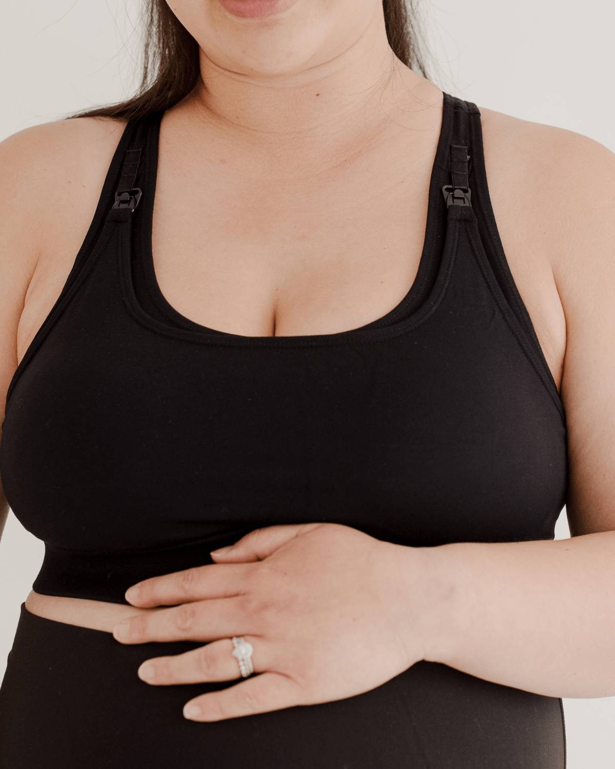 Front view of a black high-impact, racerback, maternity and nursing sports bra with one-handed clip access. Worn by a pregnant model with hands resting on her belly, standing in front of a white background. Premium maternity athletic and activewear by AXK.