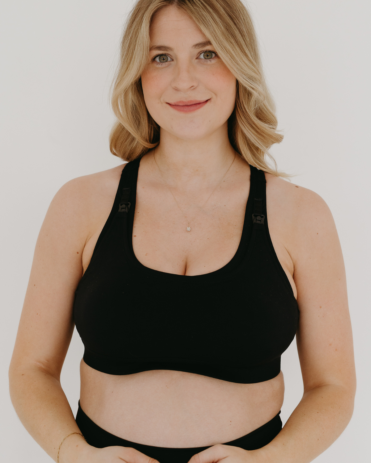 Front view of a black high-impact, racerback, maternity and nursing sports bra with one-handed clip access. Worn by a pregnant model with hands resting on her belly, standing in front of a white background. Premium maternity athletic and activewear by AXK.