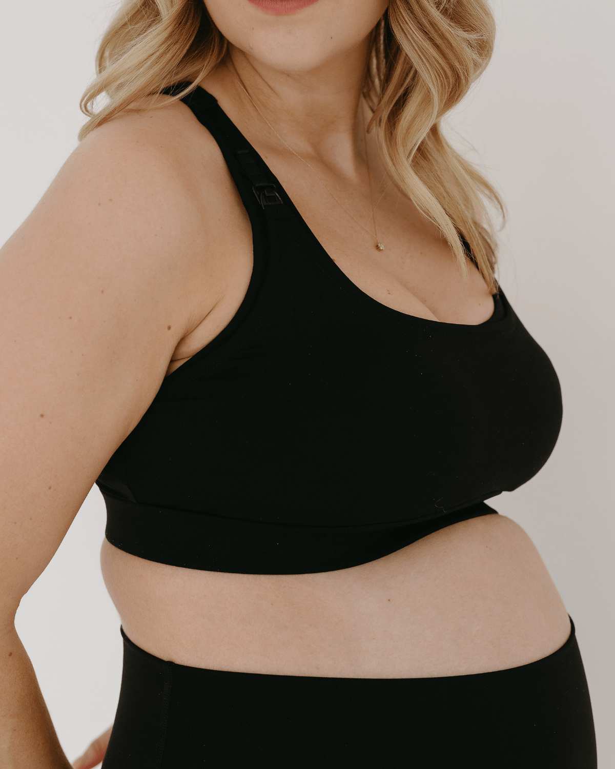 Side view of a black high-impact, racerback, maternity and nursing sports bra with one-handed clip access. Worn by a pregnant model with hands resting on her belly, standing in front of a white background. Premium maternity athletic and activewear by AXK.