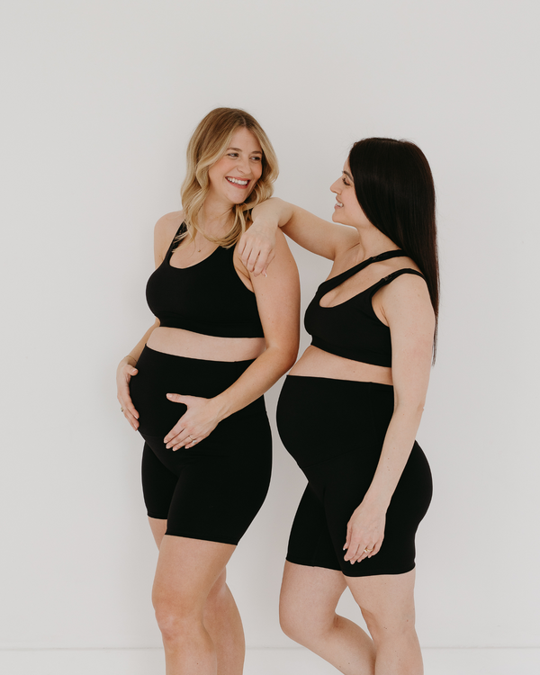 Two pregnant women wearing premium black maternity bike shorts with supportive high waist. One is wearing a one-shoulder, low-impact, nursing bra and  one is wearing a matching high-impact, racerback maternity and nursing sports bra. They are smiling at each other and standing together against a white background. Premium maternity athletic and activewear by AXK.