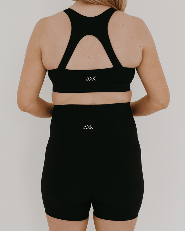 Pregnant woman wearing premium black maternity bike shorts with a supportive high waist and matching high-impact, racerback maternity and nursing sports bra, standing against a white background. Premium maternity athletic and activewear by AXK.