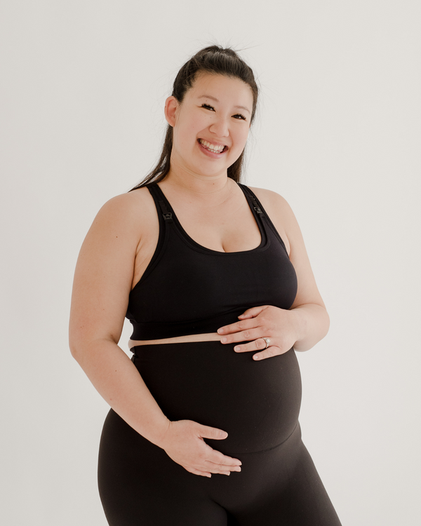 Close up photo of a pregnant woman wearing premium black maternity leggings with a supportive high waist and matching high-impact racerback maternity and nursing sports bra, she is smiling at the camera and standing against a white background. Premium maternity athletic and activewear by AXK.