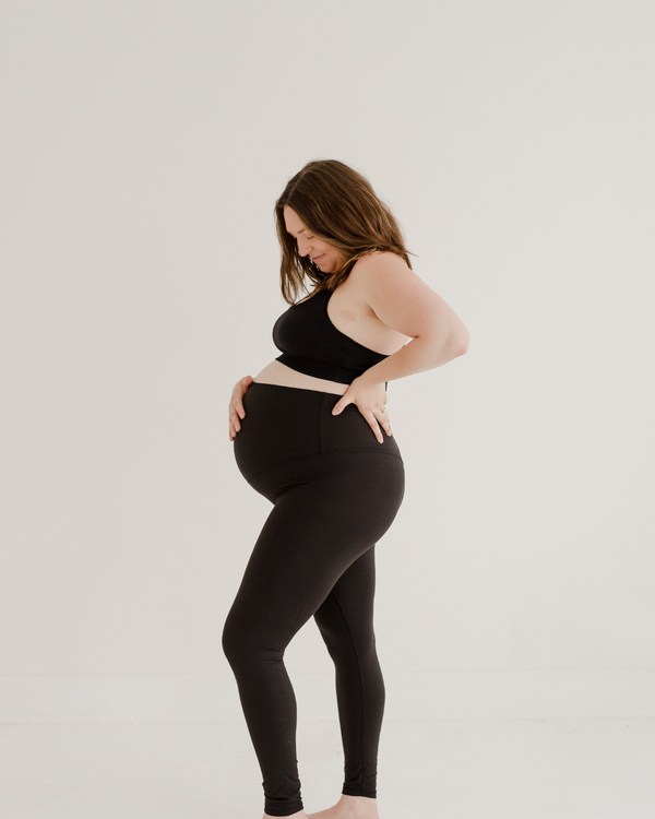Side view of a pregnant woman wearing premium black maternity leggings with a supportive high waist and matching high-impact, racerback, maternity and nursing sports bra, standing against a white background. Premium maternity athletic and activewear by AXK.
