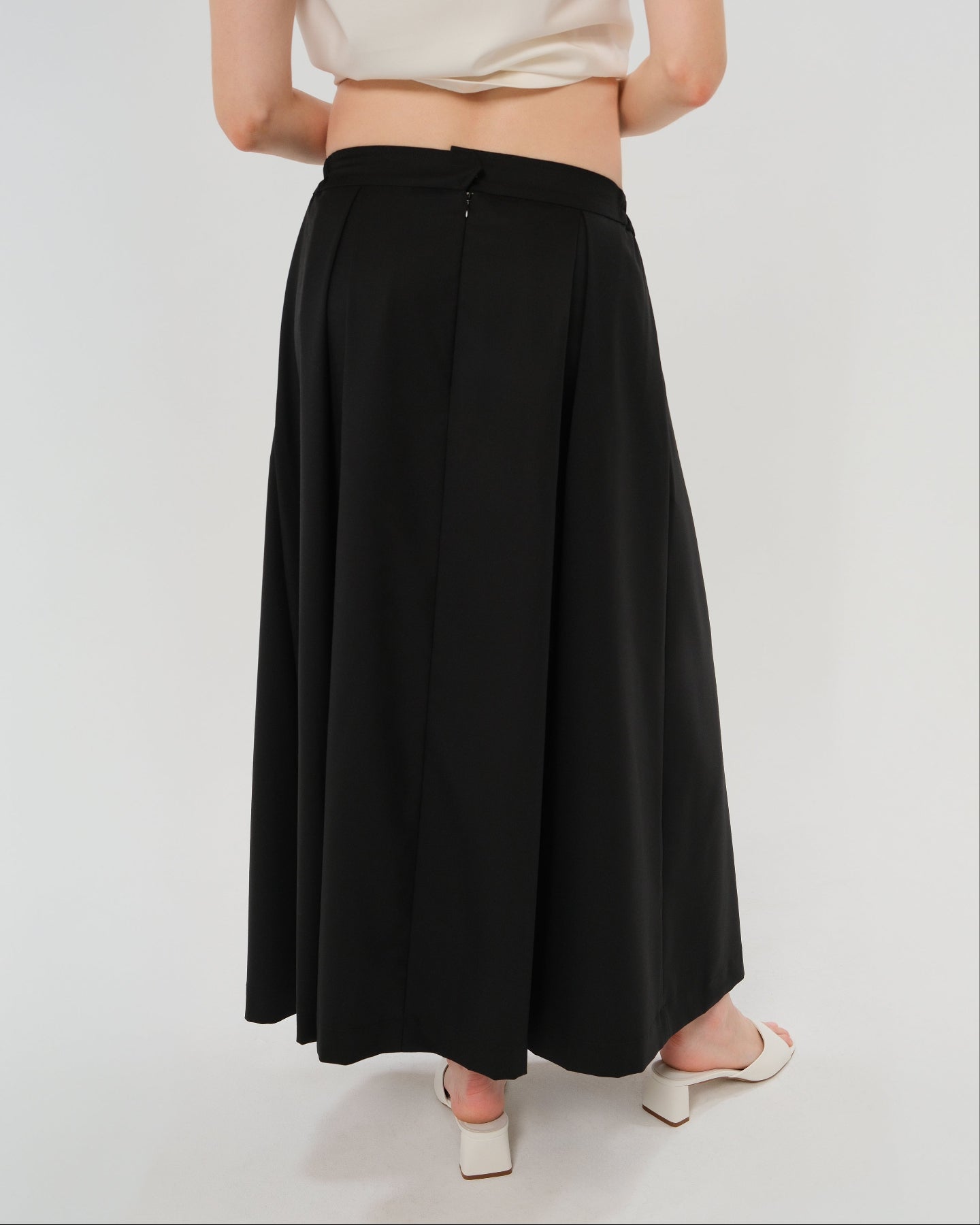 Pregnant woman wearing a black pleated designer maternity maxi maternity skirt in lightweight tropical wool by MARION. This luxury skirt can be worn over or under your belly bump. It fastens with a zip, and features deep functional pockets and a flexible elastic waistband for comfort. This refined, timeless style epitomizes quiet luxury. Perfect for maternity work wear, wedding guest dress, occasion wear, pregnancy street style, and baby shower outfits. Petite and tall friendly.