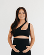 Pregnant woman wearing a premium black one-shoulder, low-impact maternity and nursing sports bra with easy clip-down access, paired with high-waist maternity leggings, standing against a light background. Premium maternity athletic and activewear by AXK.
