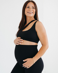 Side view of a pregnant woman wearing a premium black one-shoulder, low-impact maternity and nursing sports bra with easy clip-down access, paired with high-waist maternity leggings, standing against a light background. Premium maternity athletic and activewear by AXK.