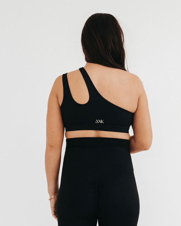 Back view of a pregnant woman wearing a premium black one-shoulder, low-impact maternity and nursing sports bra with easy clip-down access, paired with high-waist maternity leggings, standing against a light background. Premium maternity athletic and activewear by AXK.