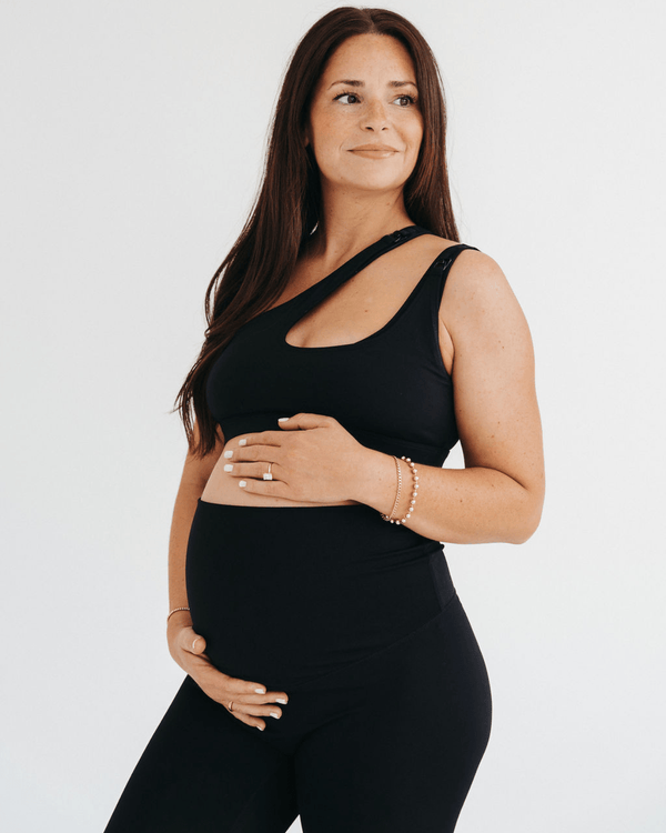 Pregnant woman wearing a premium black one-shoulder, low-impact maternity and nursing sports bra with easy clip-down access, paired with high-waist maternity leggings, standing against a light background. Premium maternity athletic and activewear by AXK.