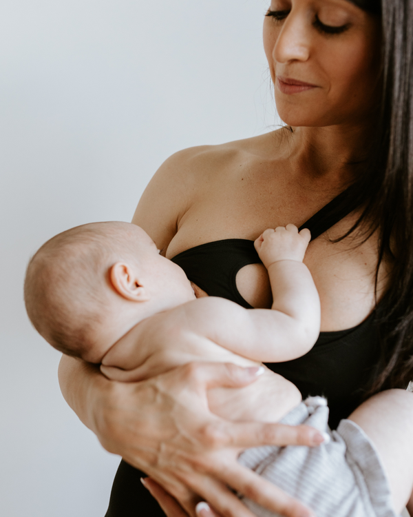 Close-up of a mother breastfeeding her baby while wearing a black one-shoulder, low-impact maternity and nursing sports bra. The bra’s pull-down nursing access is visible, showcasing functional yet stylish maternity activewear by AXK.