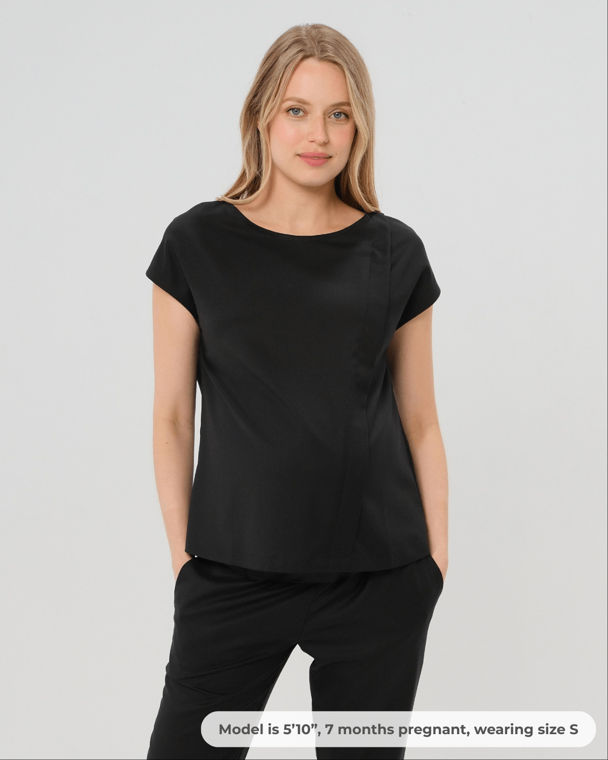 Pregnant woman wearing a black maternity short sleeve boat neck shell top with designer nursing zipper and easy, flattering elegance for work or going out. Cut in breathable, all-season tropical wool blend. This bestselling business apparel item features cap sleeves perfect for spring and summer pregnancy or breastfeeding; transitions perfectly for layering under jackets or sweaters in fall and winter. 
