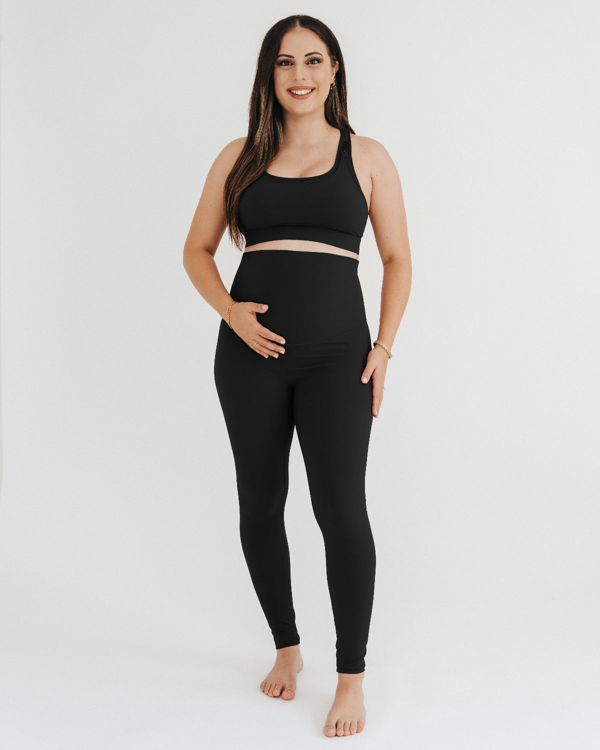 Pregnant woman wearing premium black maternity leggings with a flattering scrunch butt and matching high-impact, racerback nursing sports bra, standing against a white background. Premium maternity athletic and activewear by AXK.