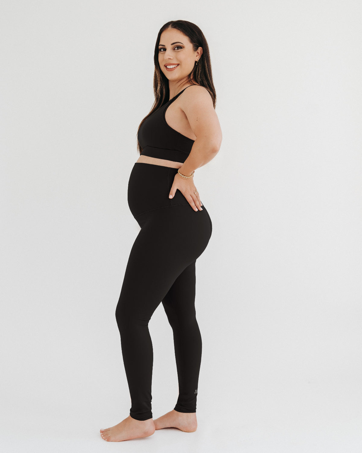Pregnant woman wearing premium black maternity leggings with a flattering scrunch butt and matching high-impact, racerback nursing sports bra, standing against a white background. Premium maternity athletic and activewear by AXK.