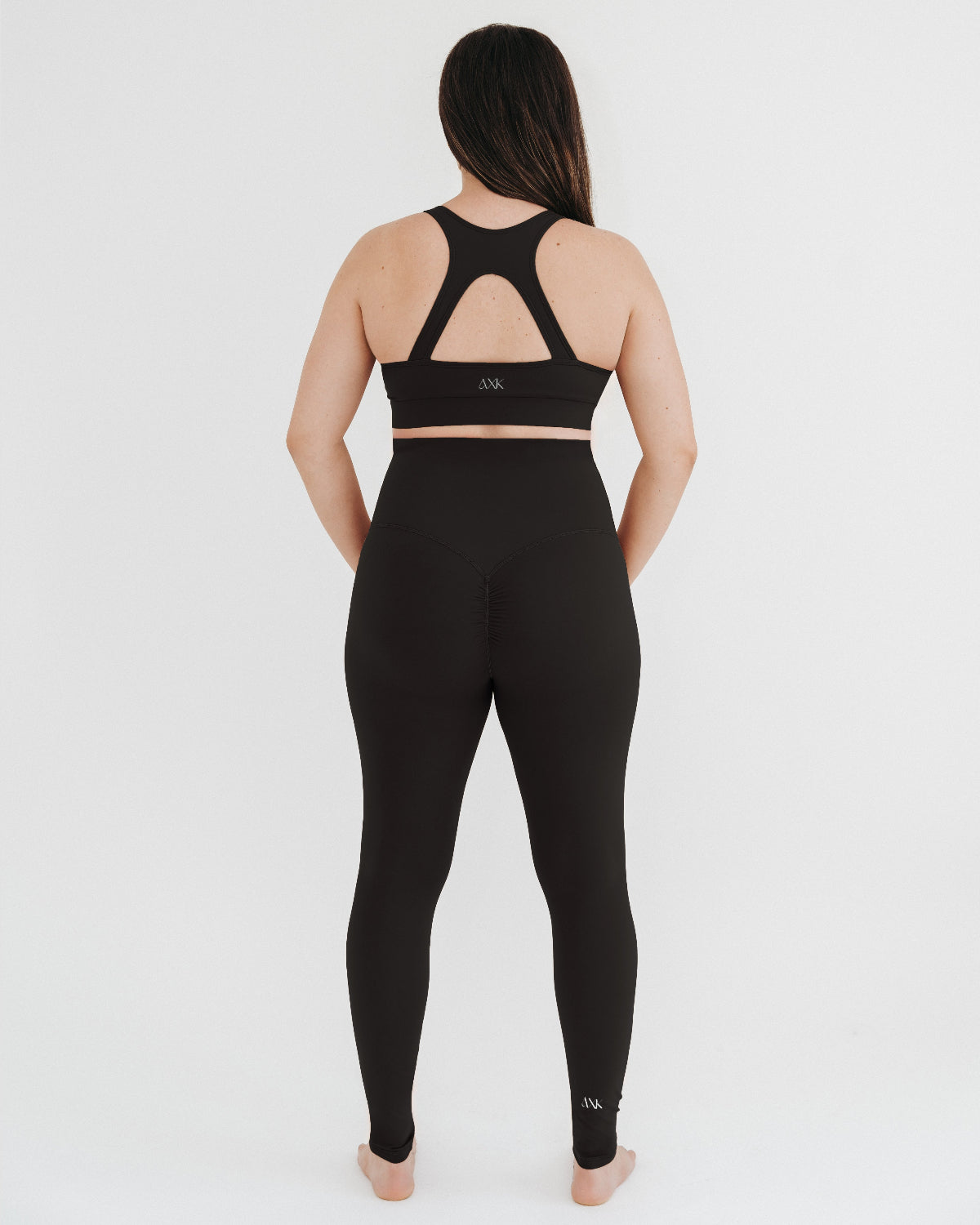 Pregnant woman wearing premium black maternity leggings with a flattering scrunch butt and matching high-impact, racerback nursing sports bra, standing against a white background. Premium maternity athletic and activewear by AXK.
