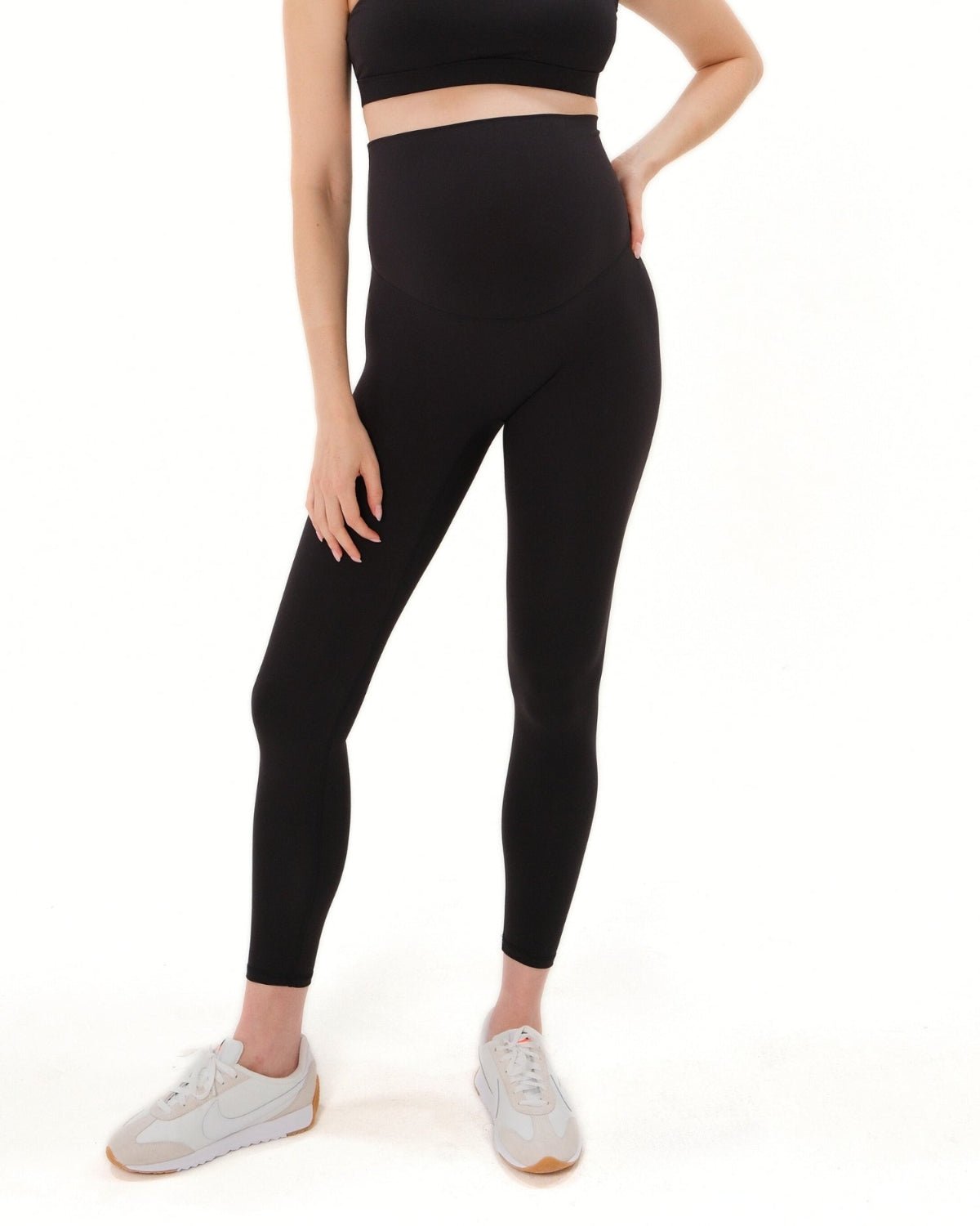 Pregnant woman wearing premium black maternity leggings with a flattering scrunch butt and matching high-impact, racerback nursing sports bra, standing against a white background. Premium maternity athletic and activewear by AXK.