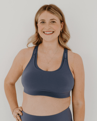 Front view of a blue high-impact, racerback, maternity and nursing sports bra with one-handed clip access. Worn by a pregnant model, standing in front of a white background. Premium maternity athletic and activewear by AXK.