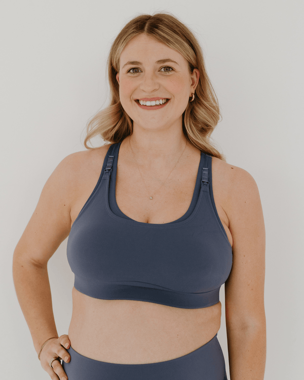 Front view of a blue high-impact, racerback, maternity and nursing sports bra with one-handed clip access. Worn by a pregnant model, standing in front of a white background. Premium maternity athletic and activewear by AXK.
