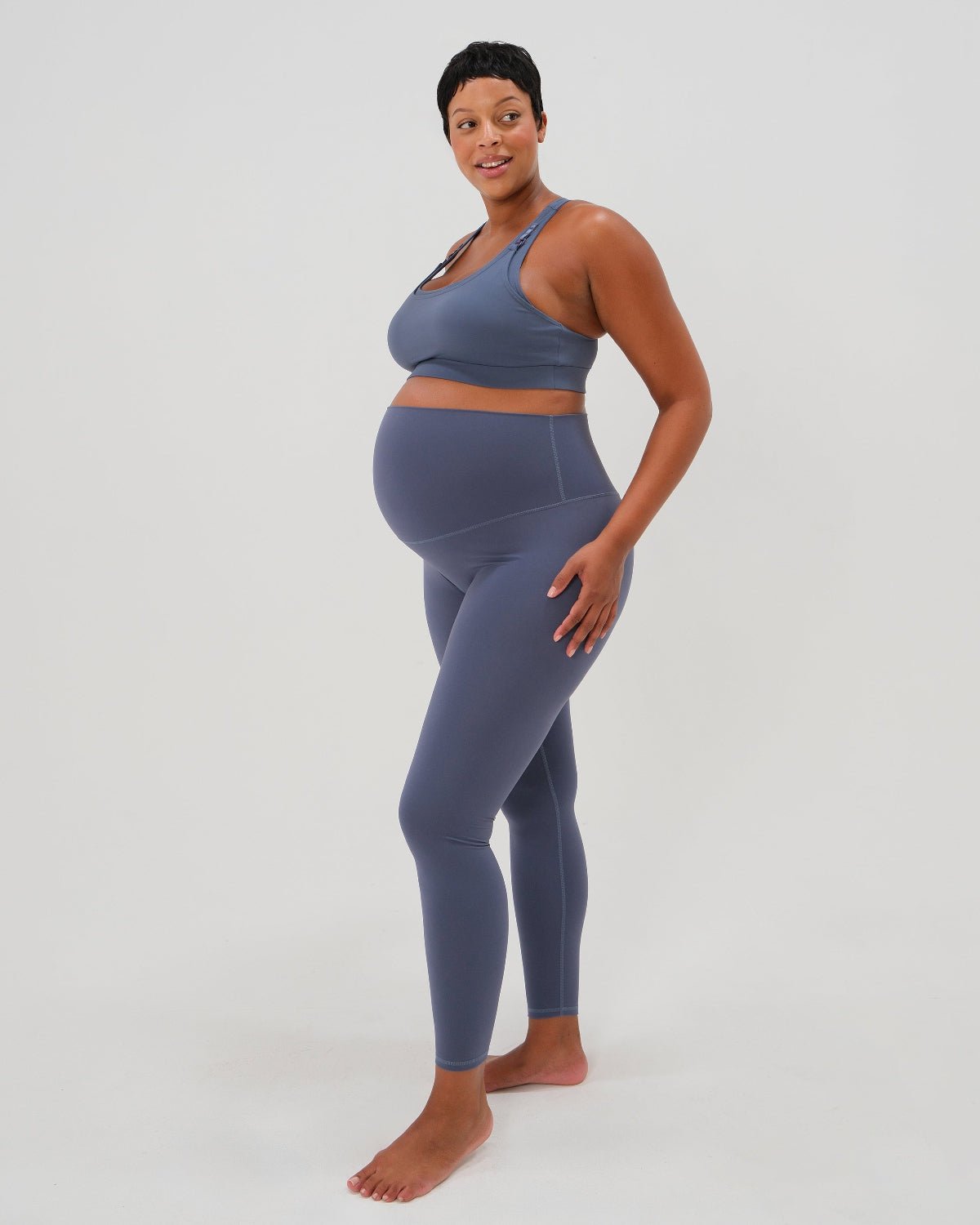 Pregnant woman wearing premium blue maternity leggings with a flattering scrunch butt and matching high-impact, racerback nursing sports bra, standing against a white background. Premium maternity athletic and activewear by AXK.