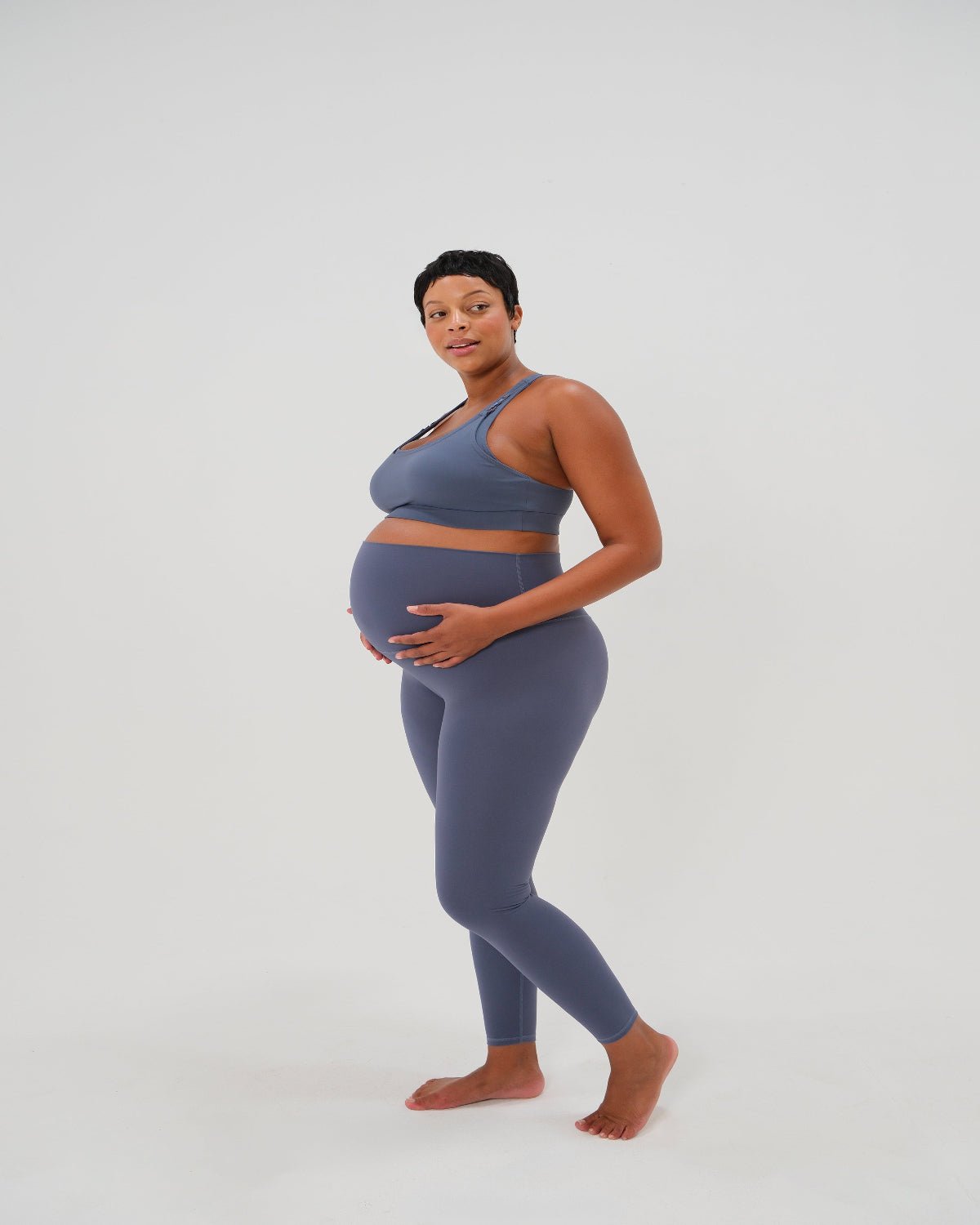 Pregnant woman wearing premium blue maternity leggings with a flattering scrunch butt and matching high-impact, racerback nursing sports bra, standing against a white background. Premium maternity athletic and activewear by AXK.
