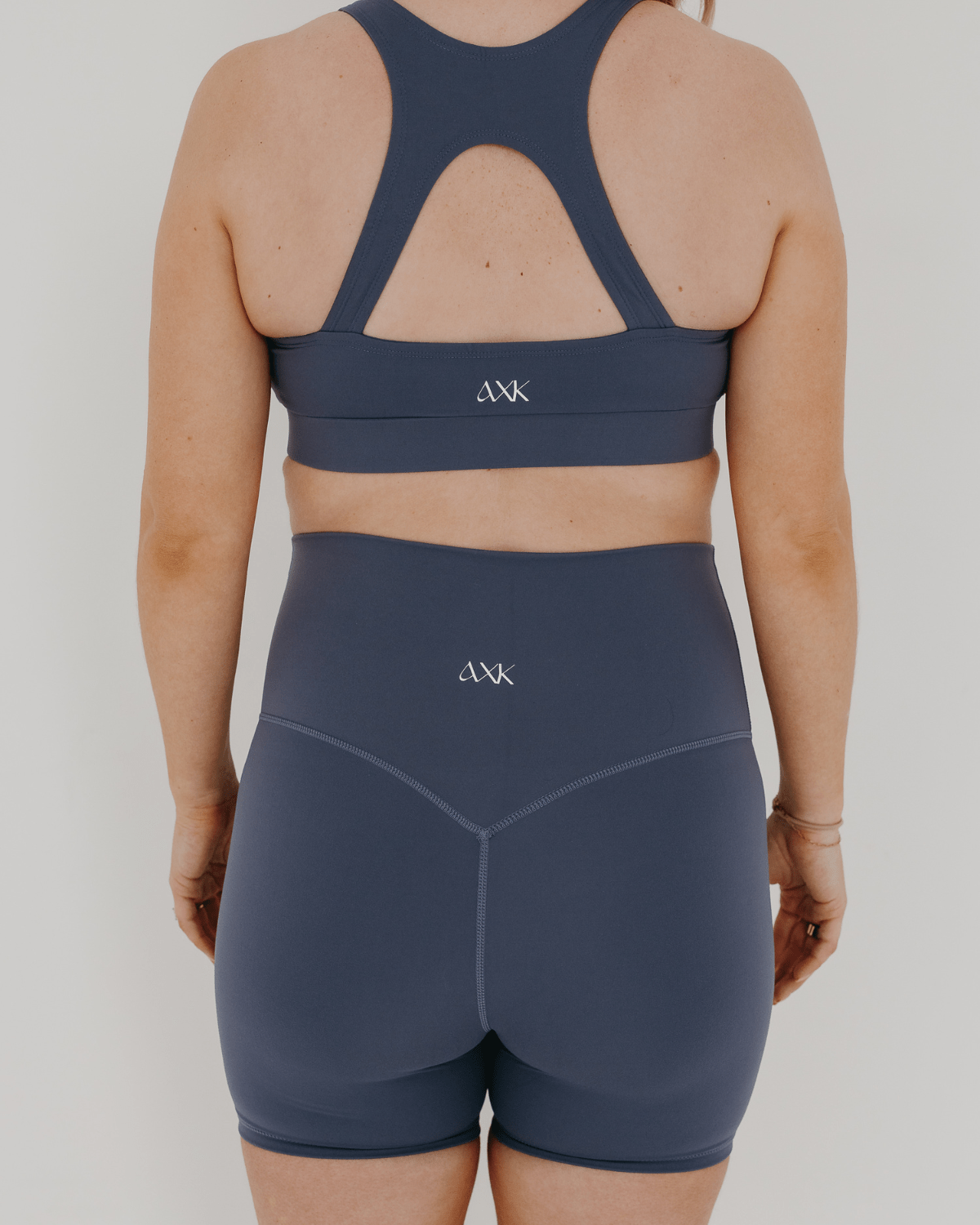 Pregnant woman wearing premium blue maternity bike shorts with a supportive high waist and matching high-impact, racerback maternity and nursing sports bra, standing against a white background. Premium maternity athletic and activewear by AXK.