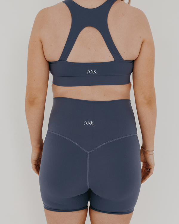 Pregnant woman wearing premium blue maternity bike shorts with a supportive high waist and matching high-impact, racerback maternity and nursing sports bra, standing against a white background. Premium maternity athletic and activewear by AXK.