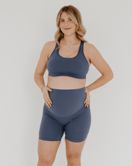 Pregnant woman wearing premium blue maternity bike shorts with a supportive high waist and matching high-impact, racerback maternity and nursing sports bra, standing against a white background. Premium maternity athletic and activewear by AXK.