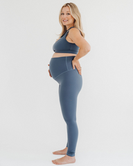 Side view of pregnant woman wearing premium blue maternity leggings with a supportive high waist and V-seam back, paired with a matching one-shoulder maternity and nursing sports bra, standing against a white background. Premium maternity athletic and activewear by AXK.