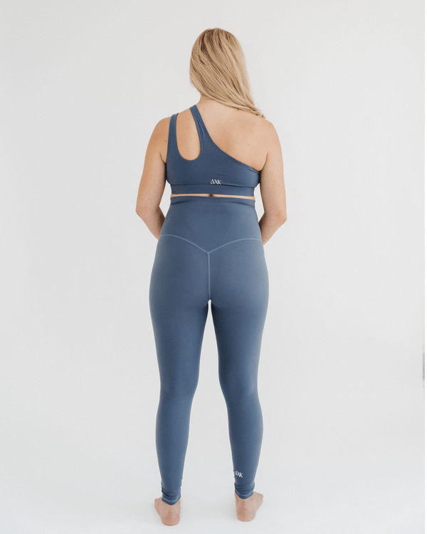 Back view of pregnant woman wearing premium blue maternity leggings with a supportive high waist and V-seam back, paired with a matching one-shoulder maternity and nursing sports bra, standing against a white background. Premium maternity athletic and activewear by AXK.