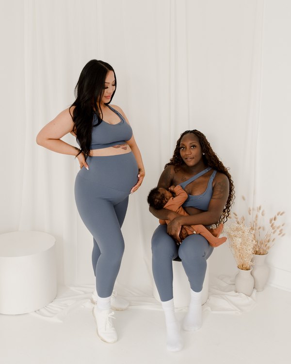 Two women wearing premium blue maternity leggings with supportive high waists and V-seam backs, paired with matching maternity and nursing sports bras. One woman is pregnant and standing, while the other is seated and nursing her baby. Both are against a white background with minimal decor. Premium maternity athletic and activewear by AXK.