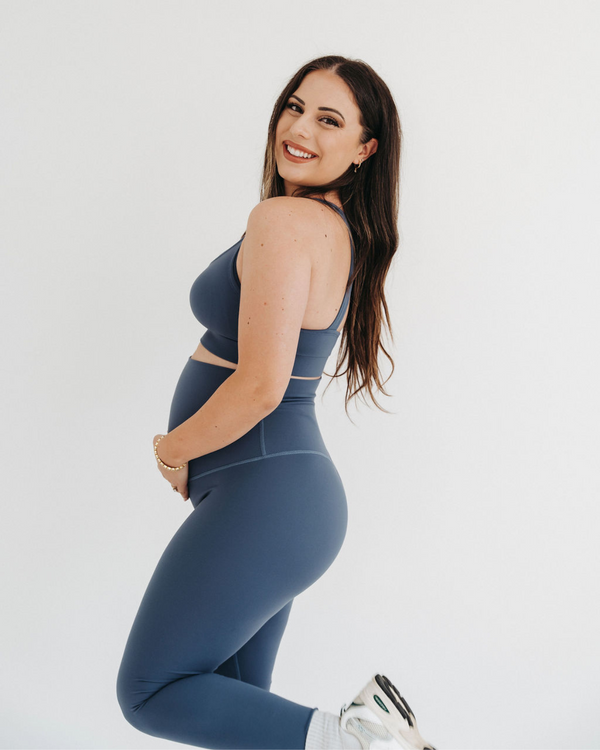 Pregnant woman wearing premium blue maternity leggings with a flattering scrunch butt and matching high-impact, racerback nursing sports bra, standing against a white background. Premium maternity athletic and activewear by AXK.