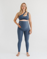 Pregnant woman wearing premium blue maternity leggings with a supportive high waist and V-seam back, paired with a matching one-shoulder maternity and nursing sports bra, standing against a white background. Premium maternity athletic and activewear by AXK.