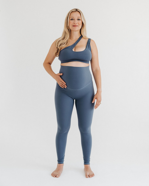 Pregnant woman wearing premium blue maternity leggings with a supportive high waist and V-seam back, paired with a matching one-shoulder maternity and nursing sports bra, standing against a white background. Premium maternity athletic and activewear by AXK.