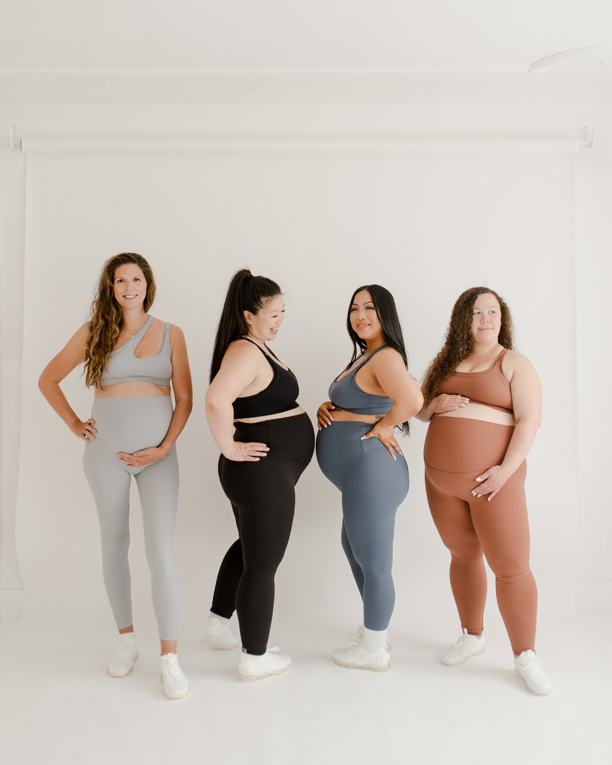 Four pregnant women wearing premium AXK maternity leggings with supportive high waists and V-seam backs, paired with matching maternity and nursing sports bras. Each woman wears a different color set. One in light grey, black, blue, and brown standing together against a white background. Premium maternity athletic and activewear by AXK.